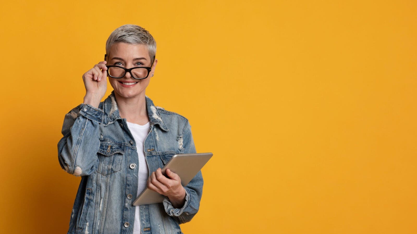 Modern Teacher. Portrait Of Stylish Mature Woman In Eyeglasses Holding Digital Tablet And Looking At Camera, Posing On Yellow Background, Panorama
