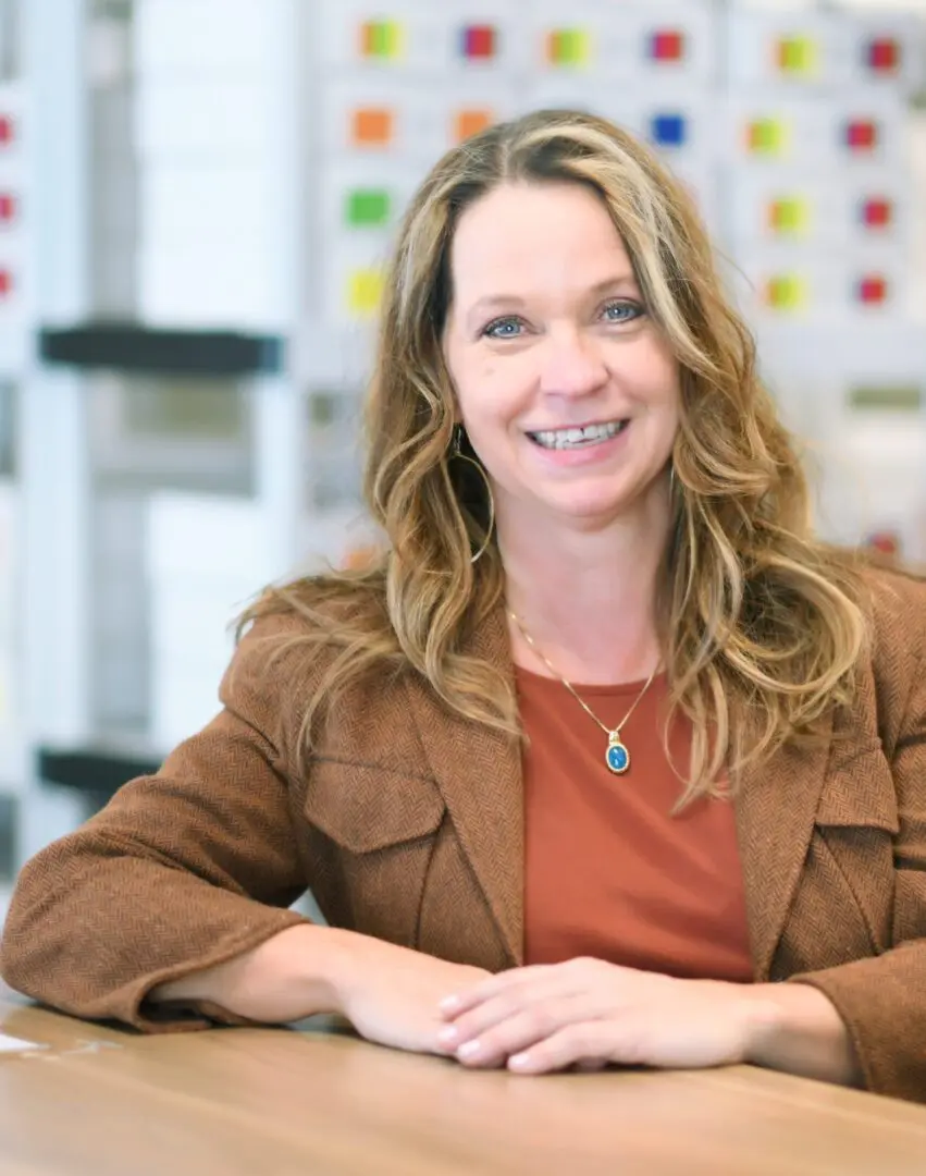 A smiling woman with long hair, wearing a brown blazer, seated at a table in a brightly lit office.