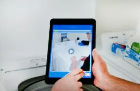 A person using a tablet to control a washing machine, displayed with video instructions on the screen.