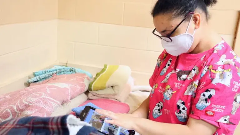 A healthcare worker in a pink uniform and mask using a tablet in a room with a simple bed and towels.