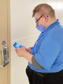 Man in blue shirt and mask using smartphone near a digital keypad on a wall.