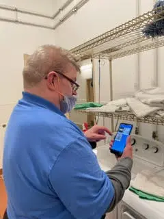 A man in a blue shirt and glasses, wearing a mask, uses a smartphone in a room with shelving and linens.