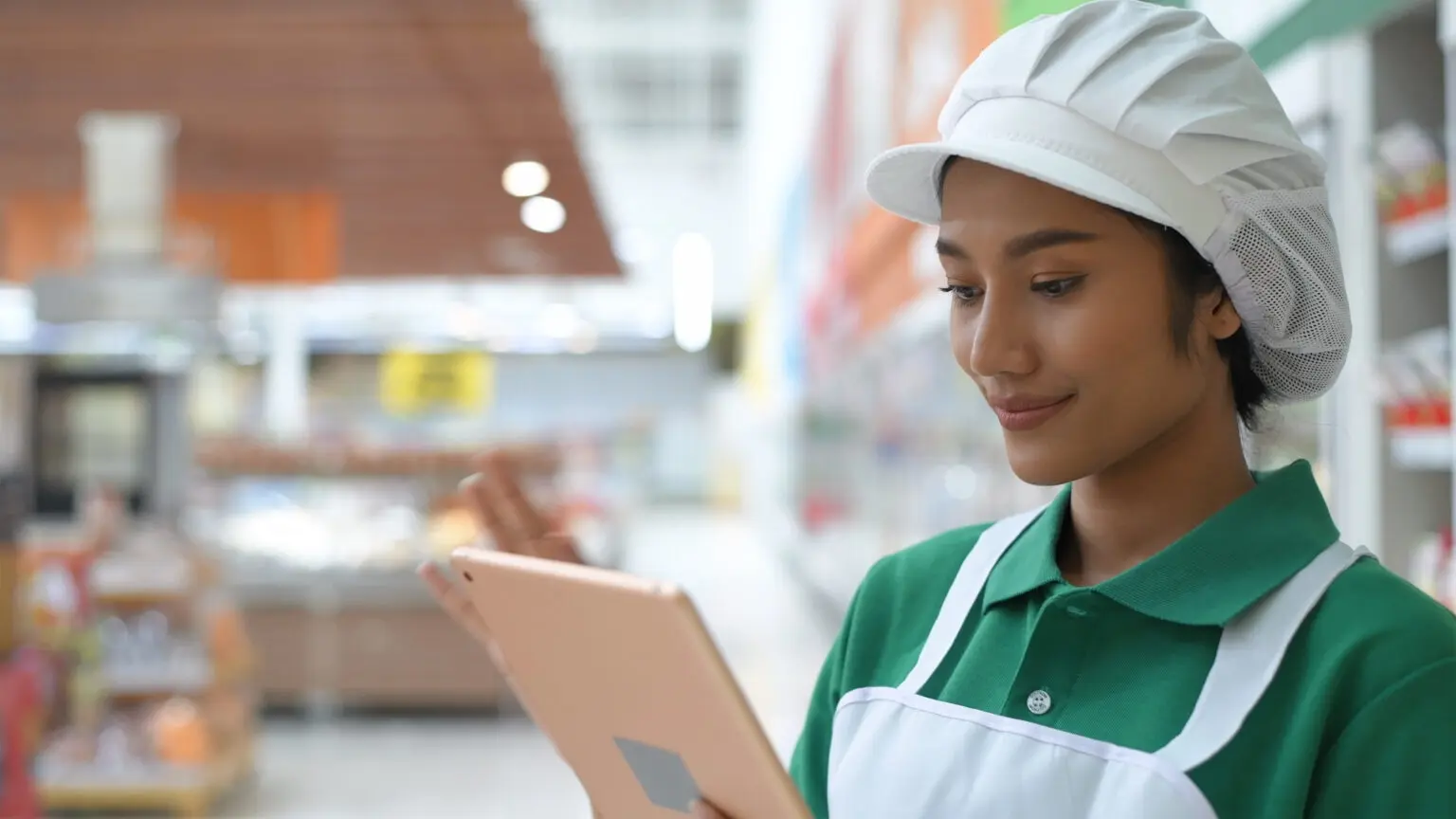 A supermarket employee in a green uniform and white hat using a tablet inside the store.