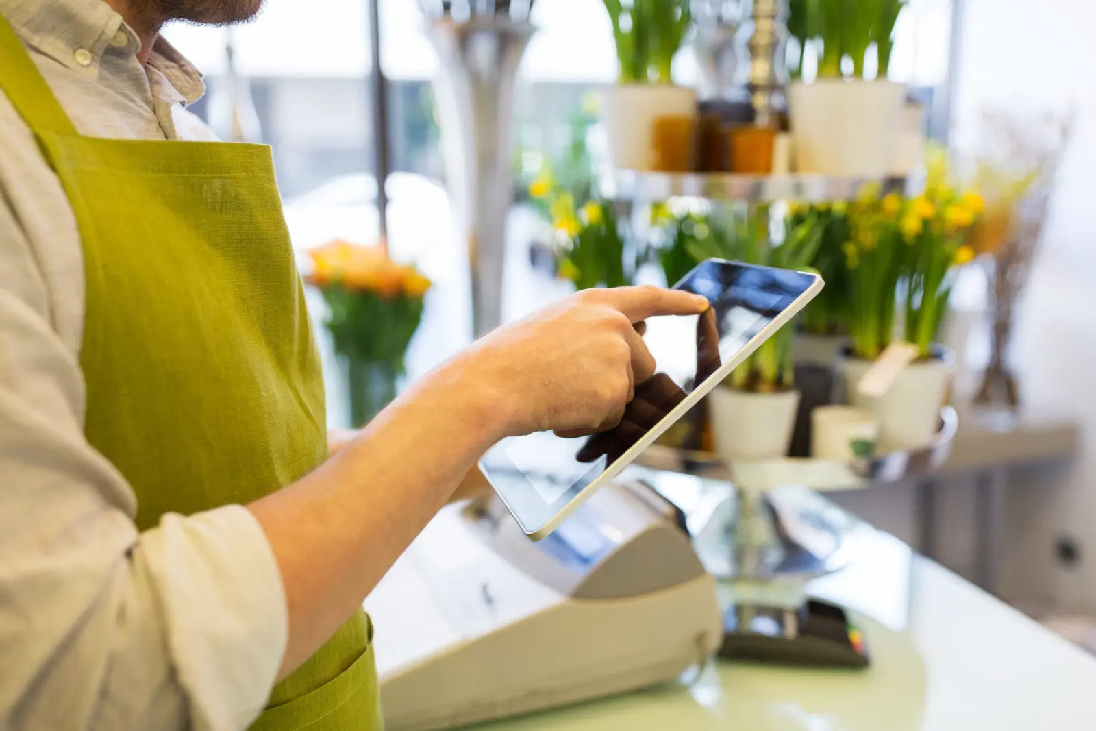 A male barista in a green apron uses a tablet at the counter of a bright cafe.