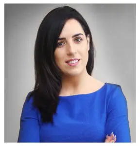 Portrait of a woman with shoulder-length dark hair wearing a blue top, arms crossed, against a gray background.