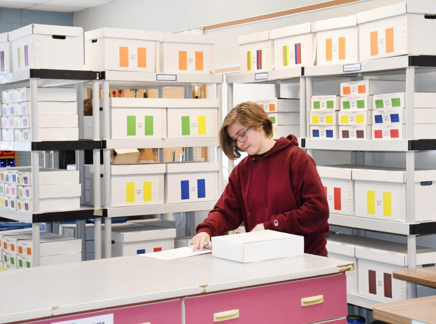 A woman in red shirt holding box near shelves.