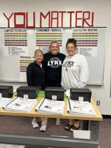Three women standing in front of a table with laptops.