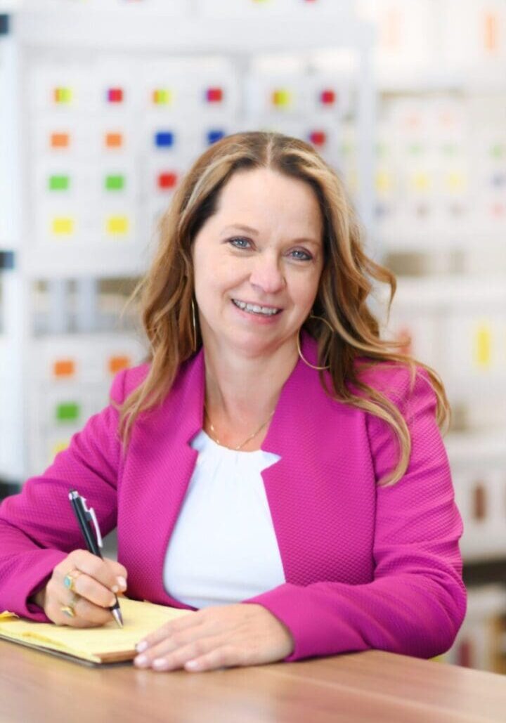 A businesswoman in a pink blazer smiles while writing in a notebook at a table, with a colorful file cabinet in the background.